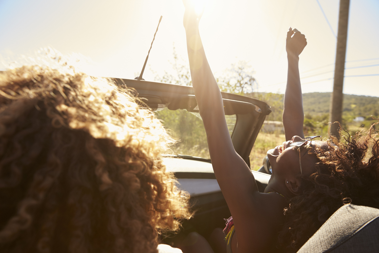 Built By She_Taking Risks_Young couple in an open top car, woman with arms raised