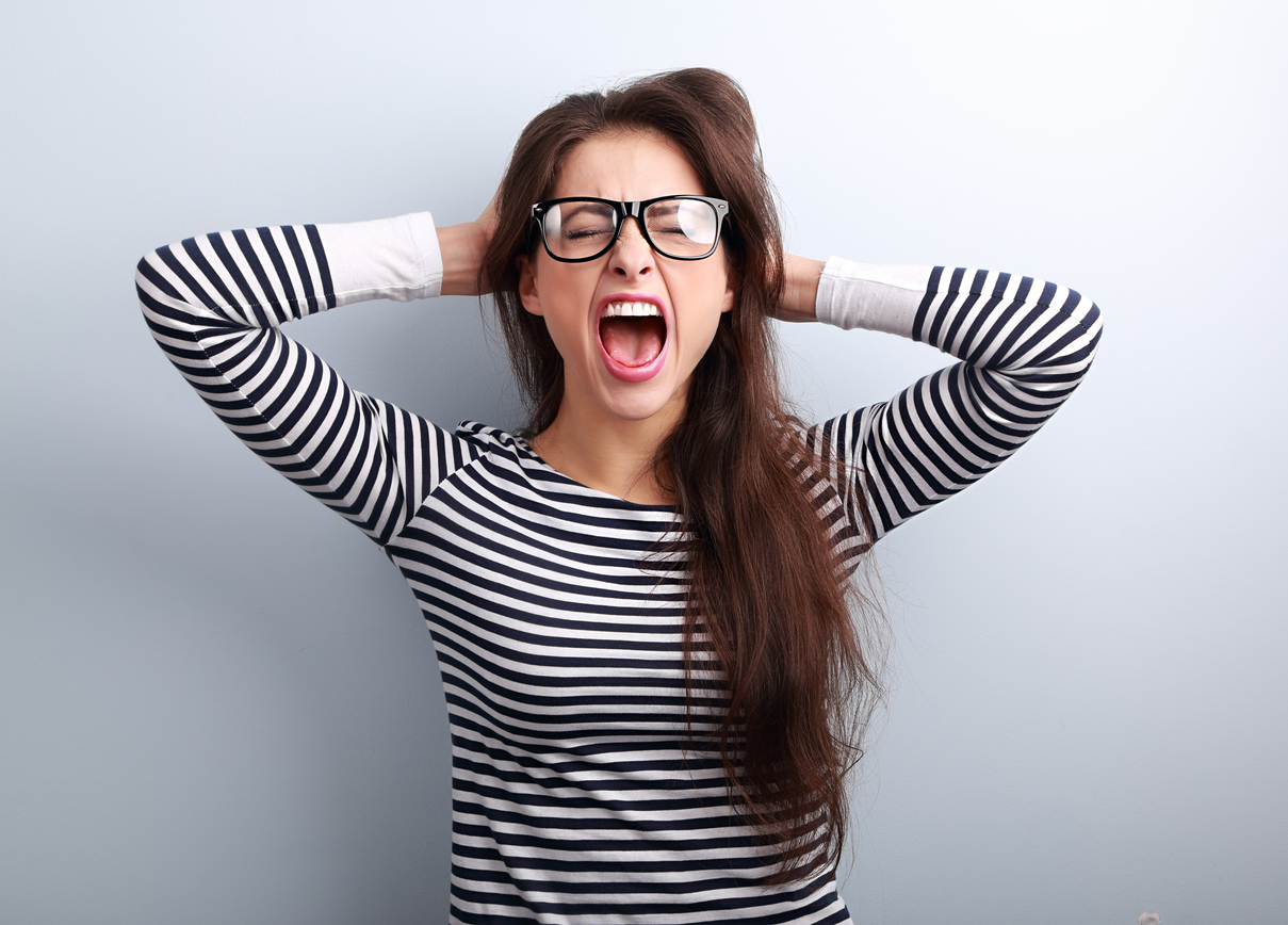 Built By She - Anger young business woman in glasses strong screaming with wild open mouth and holding head the hands on blue background