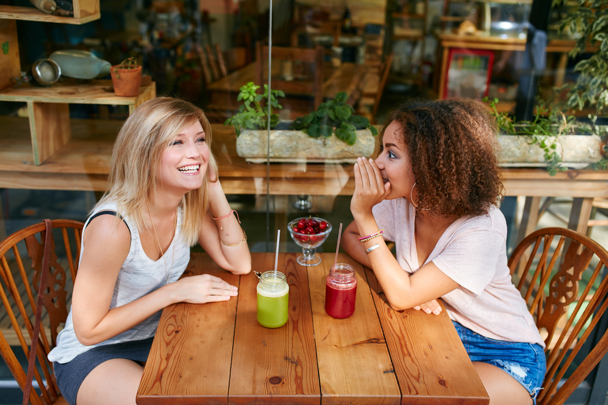 Built By She - Young female friends having private gossips at sidewalk cafe. Two young women gossiping and whispering outdoors in a coffee shop.