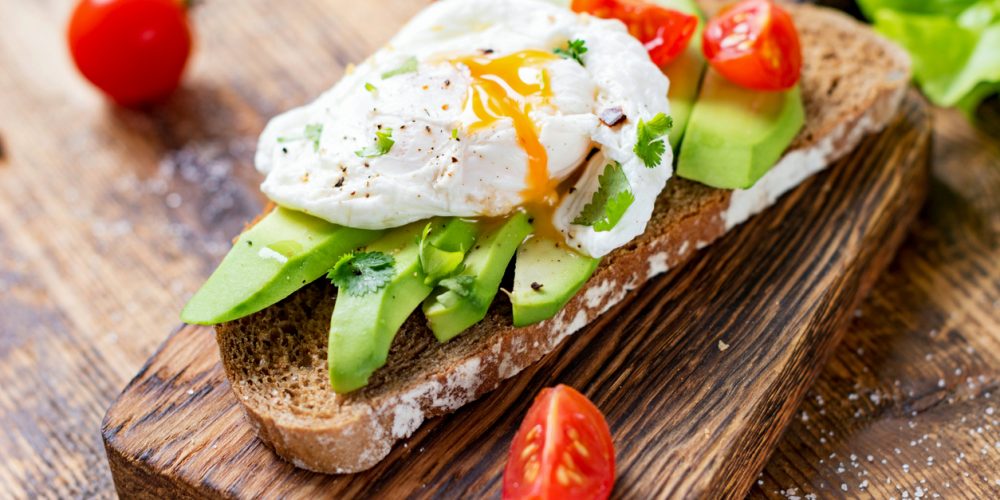 Built By She - Tasty sandwich with avocado, tomato and poached egg on wooden chopping board, close up, selective focus. Healthy delicious breakfast or lunch