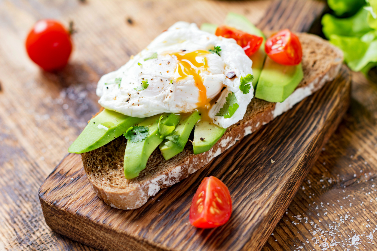 Built By She - Tasty sandwich with avocado, tomato and poached egg on wooden chopping board, close up, selective focus. Healthy delicious breakfast or lunch